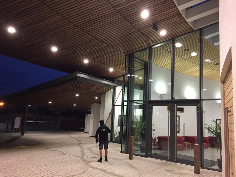 Cleaner working on tall glazing at a lit commercial entrance in the evening